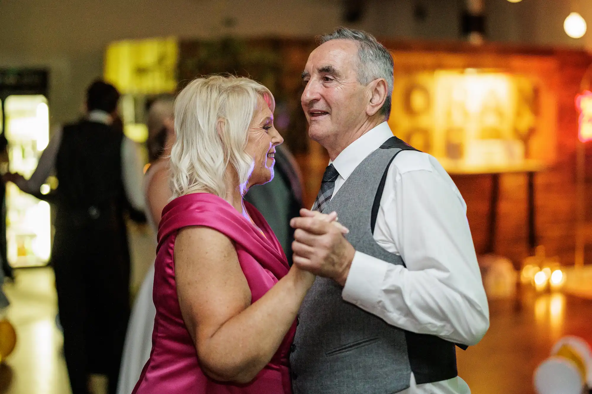 An elderly couple smiles and dances together at an indoor event. The woman wears a bright pink dress, and the man is dressed in a white shirt, tie, and grey waistcoat. Soft ambient lighting creates a warm, festive atmosphere in the background.