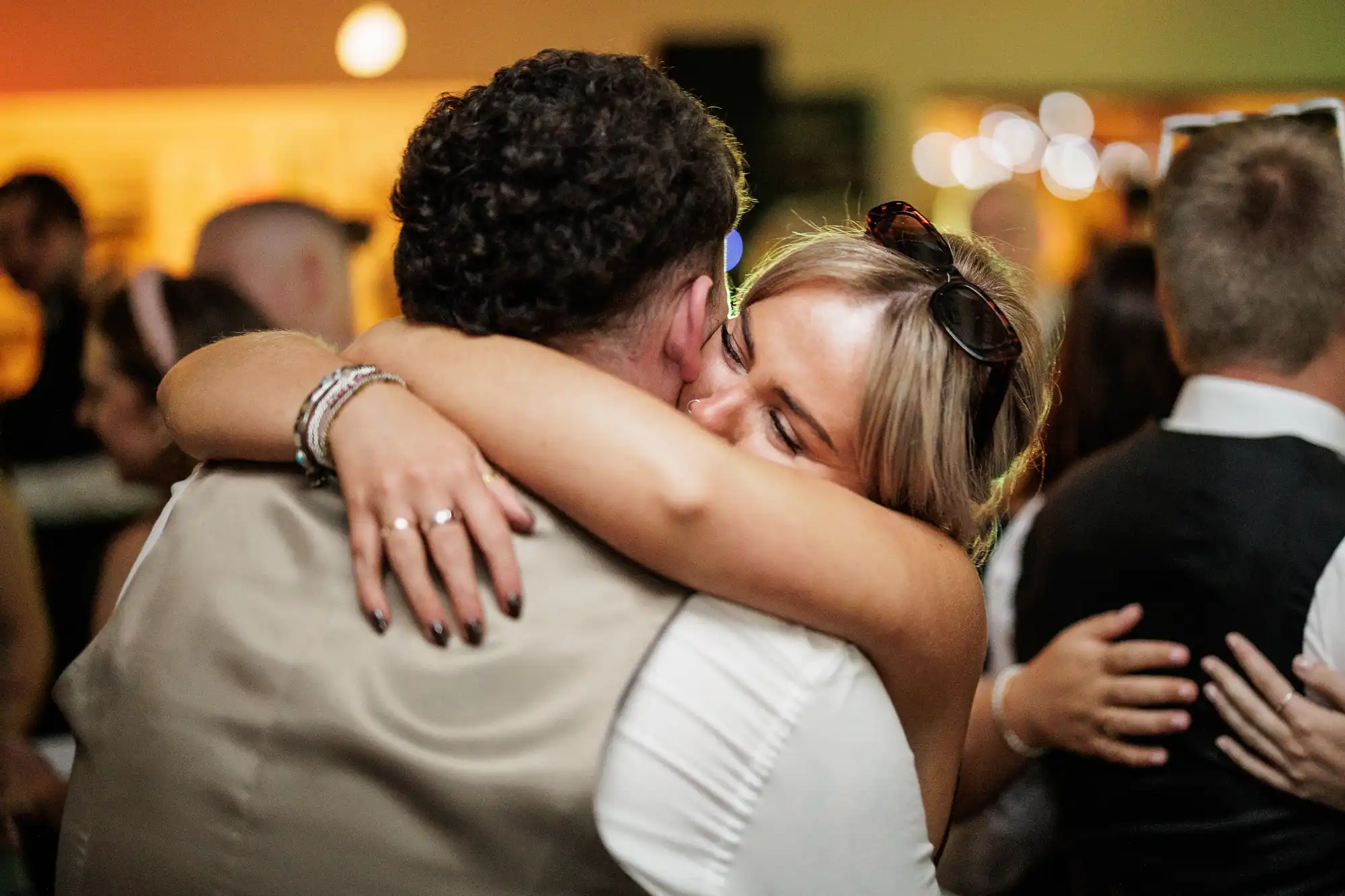 A blonde woman wearing sunglasses on her head embraces a man in a tan waistcoat, smiling with her eyes closed. They are surrounded by other people dancing in a warmly lit indoor setting, suggesting a celebratory event.