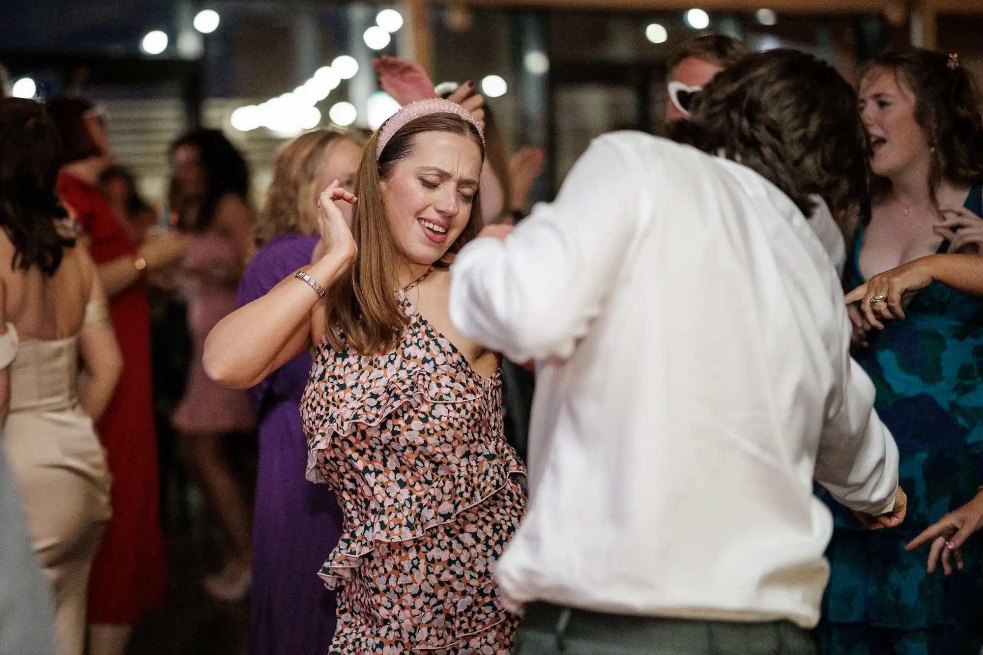 A woman in a pink patterned dress and headband dances with her eyes closed at a lively indoor event. A man in a white shirt dances in front of her. Other guests in colourful outfits are visible in the background, enjoying the festive atmosphere.