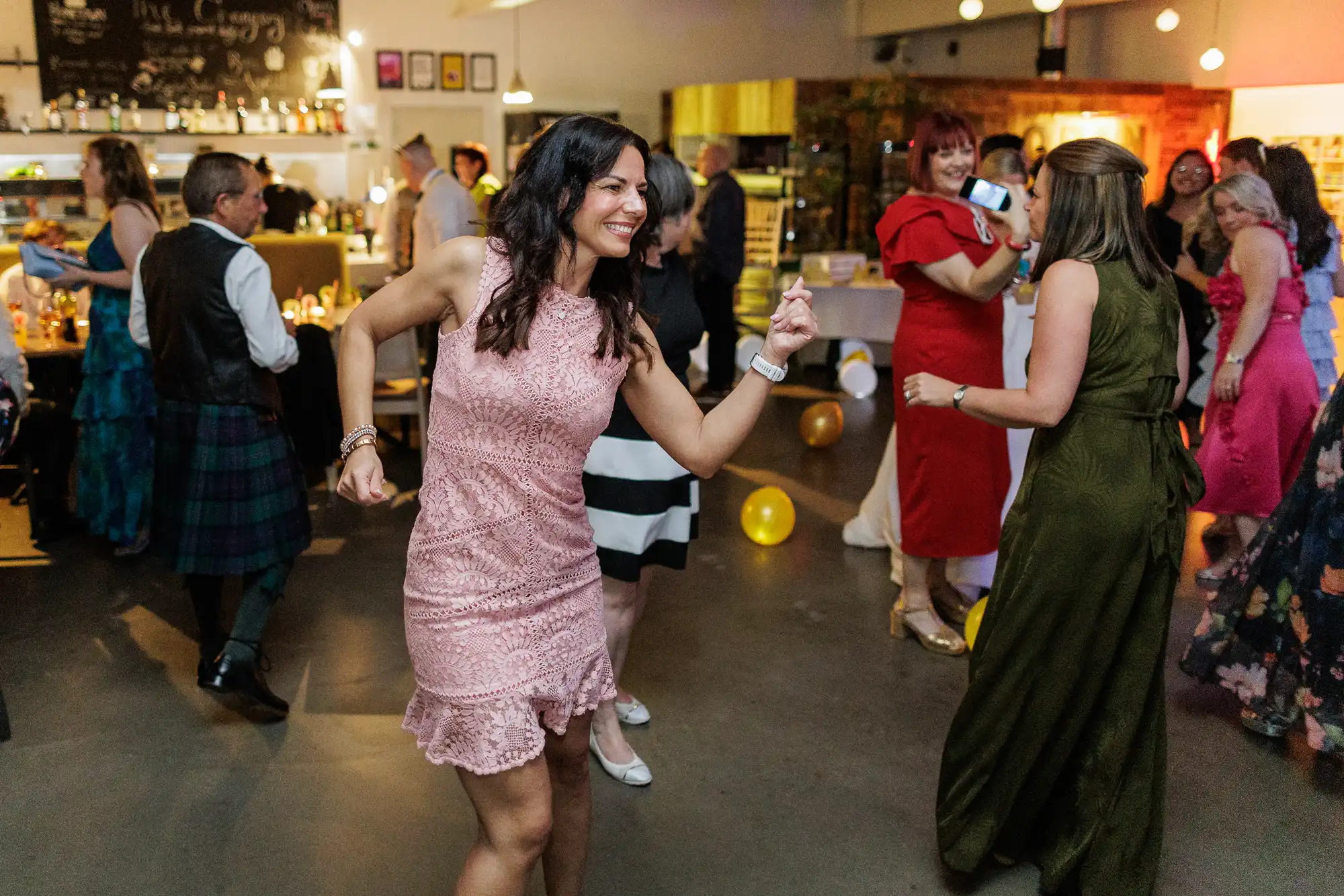 A woman in a pink lace dress smiles and dances at a lively indoor party. Other guests, dressed in colourful outfits, chat, dance, and take photos. The decorated venue glows with warm lighting and balloons scattered on the floor.