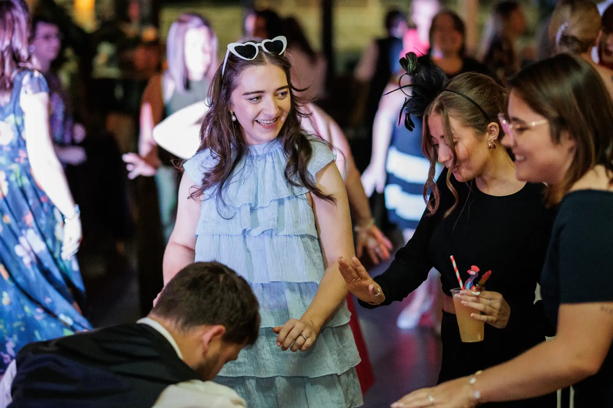 A group of people are dancing at a lively indoor event. A woman in a frilly light blue dress with heart-shaped sunglasses on her head smiles whilst dancing with others. Some people hold drinks, and the background is filled with more dancers.