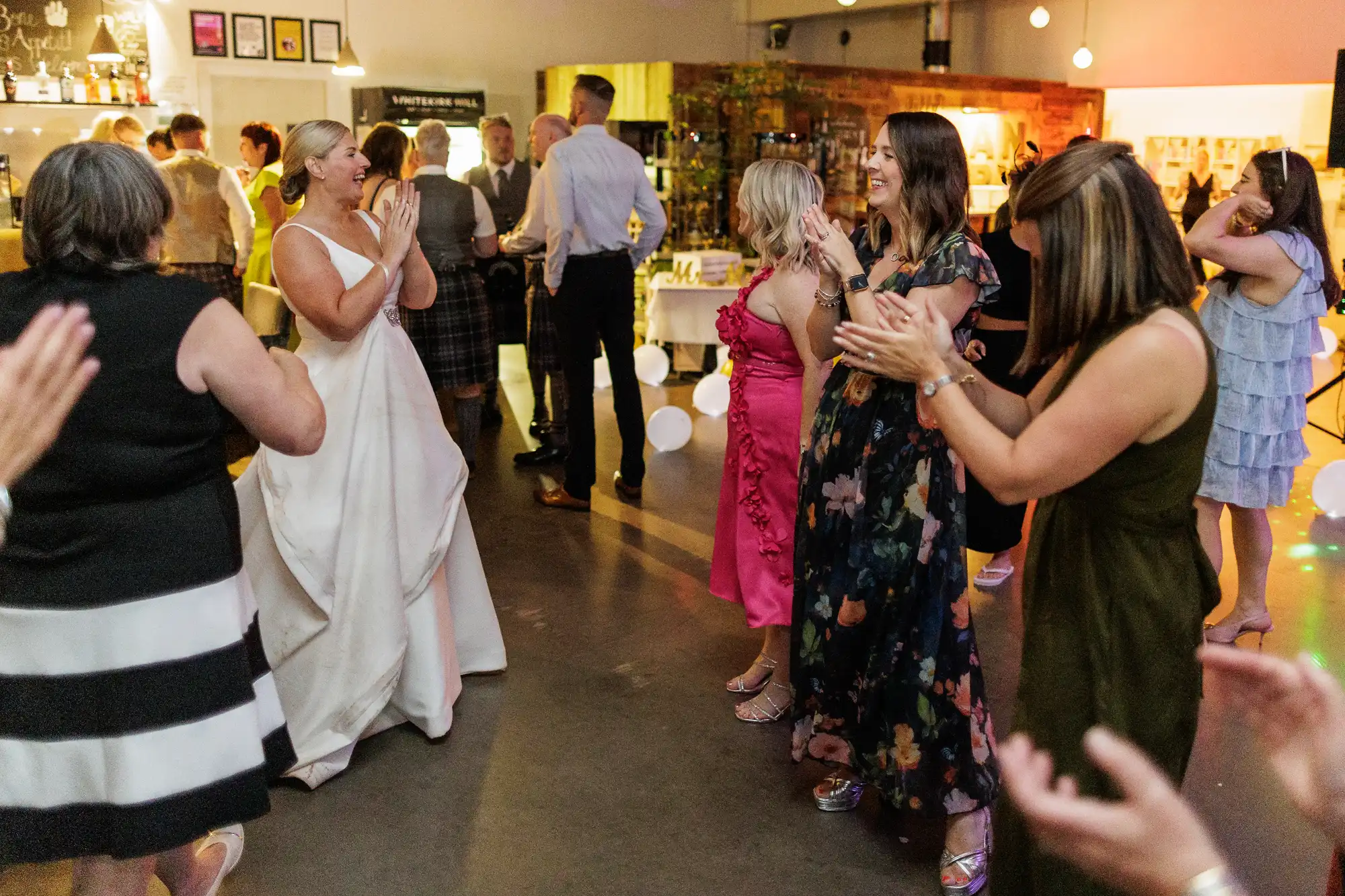 A bride in a white dress joyfully claps whilst dancing with guests at a wedding reception. Other people, dressed in colourful dresses and formal attire, clap and smile on a warmly lit dance floor with balloons and festive decorations in the background.